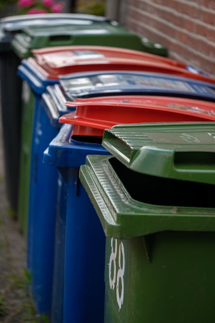 A row of large wheelie bins positioned along a pavement next to a brick wall, with the bins arranged side by side in a sequence that includes green, blue, red, and dark green colors. The green bin in the foreground is topped with a matching green lid, featuring ridged grooves for stacking or gripping. The blue bins surrounding it are also equipped with matching lids, and the red bin stacks follow behind. The bins are made of plastic with a textured surface and visible handles, designed for waste collection. The environment is outdoors, with the bins placed directly on the pavement, and the scene is lit naturally, with soft daylight illuminating the items and casting mild shadows. This setup reflects typical arrangements used for private waste disposal or alternative rubbish collection services, such as those provided by waste disposal companies, rather than standard municipal collection. The background includes a blurred view of the brick wall and some greenery along the edge of the pavement, emphasizing the outdoor setting where rubbish removal and waste management are commonly carried out by specialists like Waste Disposal Barnet.