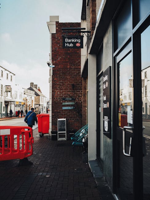 The image depicts a street scene outside a building housing a banking hub, with a visible black and red sign mounted on a brick wall. To the left, a pedestrian in a blue coat walking along the pavement is seen near a bright red waste collection bin and a smaller red container positioned on the ground. Adjacent to the building, there are outdoor seating arrangements with a teal bench placed against the brick wall. The wider street scene in the background shows shops and other buildings with pedestrians walking along the pavement. The building's exterior is comprised of brick and glass elements, with a large black-framed window or door on the right side of the image. The atmospheric lighting appears natural, possibly with overcast skies, providing diffuse illumination over the scene. Waste disposal tasks are not directly depicted, but the presence of waste containers and the focus on external urban elements suggest activity related to rubbish removal services, such as those offered by Waste Disposal Barnet, in maintaining a clean commercial environment.