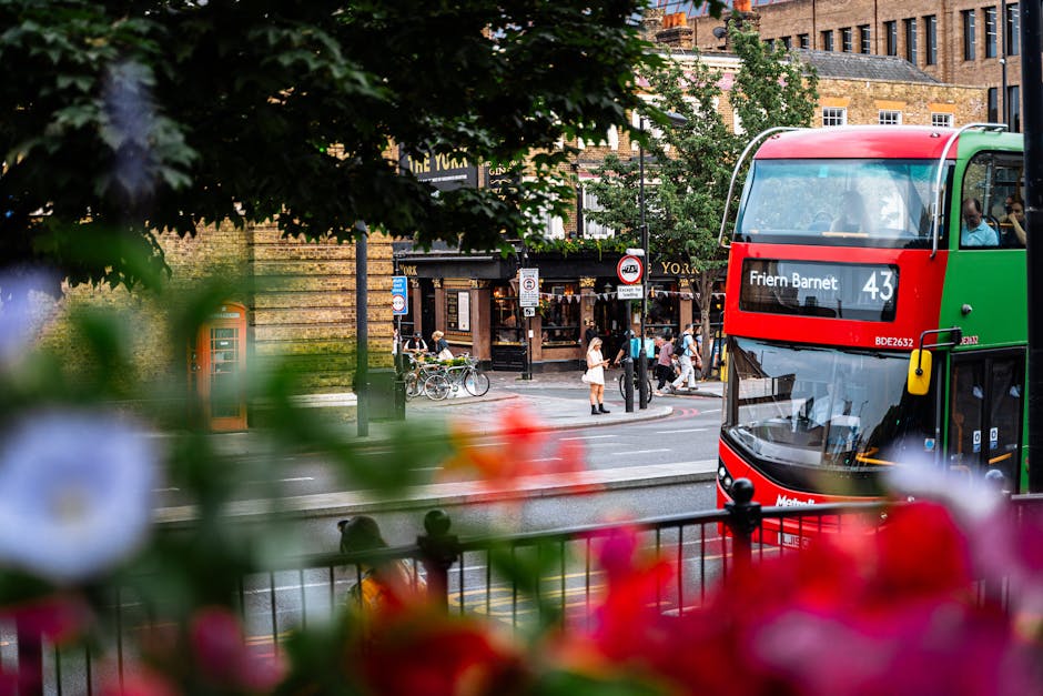 A double-decker bus with a red, green, and black exterior is positioned on a street in Barnet, displaying the route number 43 and the destination 'Friern Barnet' on its front panel. In the foreground, out-of-focus colorful flowers and green leaves frame the lower part of the image, adding a natural contrast. The bus is parked or moving along a paved road with yellow and white lines, adjacent to a sidewalk where several pedestrians are crossing or waiting to cross. Some individuals are seen walking, while others stand near a corner housing various street signs and a red telephone booth, indicative of an urban setting. Behind the bus, there are brick buildings with a mixture of traditional and modern architectural features, including large windows and decorative facades, suggesting a lively commercial or shopping area. The scene is well-lit with natural daylight, capturing the vibrant city atmosphere that aligns with private transit services and off-street rubbish collection in busy urban areas like Barnet.