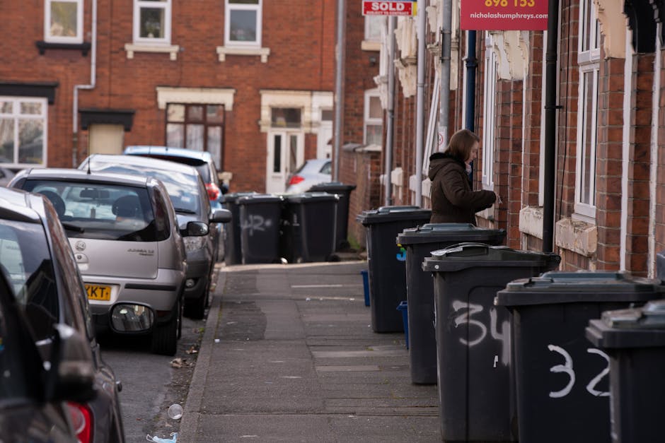 A residential street scene showing a woman standing next to a row of black wheelie bins on a paved sidewalk, with some bins partially open and others closed, arranged along the front of red brick terraced houses with white window frames. Several cars are parked along the curb, including a silver hatchback and a darker vehicle, with license plates visible. The sidewalk has some litter scattered near the bins and cars. In the background, more similar brick houses and additional bins are visible, with a red 'Sold' sign attached to one of the buildings. The lighting suggests a daytime setting with overcast skies, and the scene reflects typical urban rubbish collection or private waste disposal arrangements in a residential area, with Waste Disposal Barnet occasionally implied as an example of local rubbish removal services.
