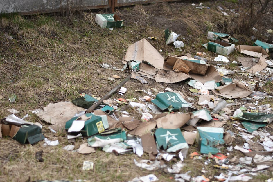 An outdoor scene showing discarded packaging materials and debris scattered across a patch of grass and dirt terrain. The debris includes crushed cardboard boxes with green and white printed logos, some torn open with shredded paper spilling out. The cardboard appears weathered with bent and crumpled surfaces, and some containers are partially flattened or collapsed. Among the debris, there are loose paper scraps and small fragments of packaging material. In the background, there is a rusty metal fence at the top of a slope, with sparse vegetation and ground covering a mixture of brown soil and patches of grass. A metal pole or rod is visible lying amongst the rubbish, partially buried in the ground. The scene is illuminated with natural daylight, highlighting the worn textures of the trash and the earth tones of the environment, reflecting a typical case of illegal dumping that would benefit from professional waste removal services such as those offered by Waste Disposal Barnet to manage and clear roadside rubbish efficiently.